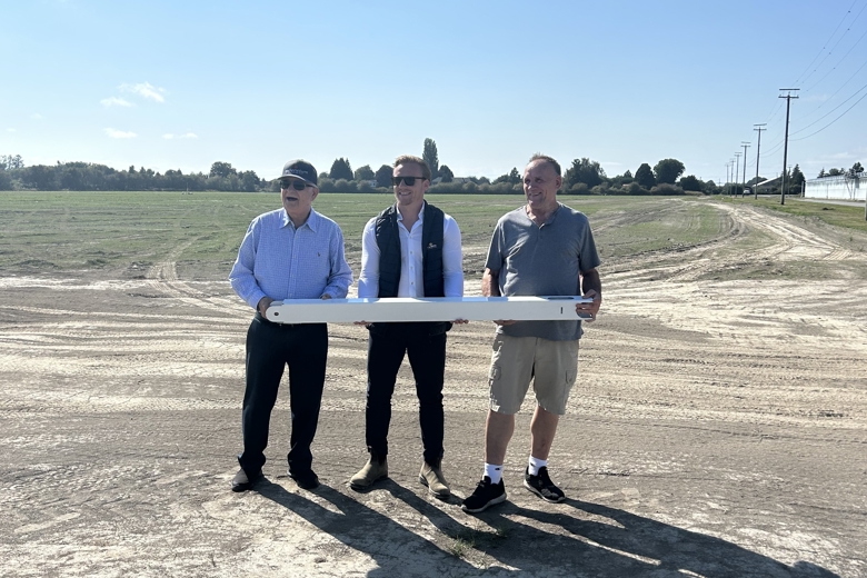 Drie mannen staan op een open bouwterrein en houden gezamenlijk een lang wit object vast